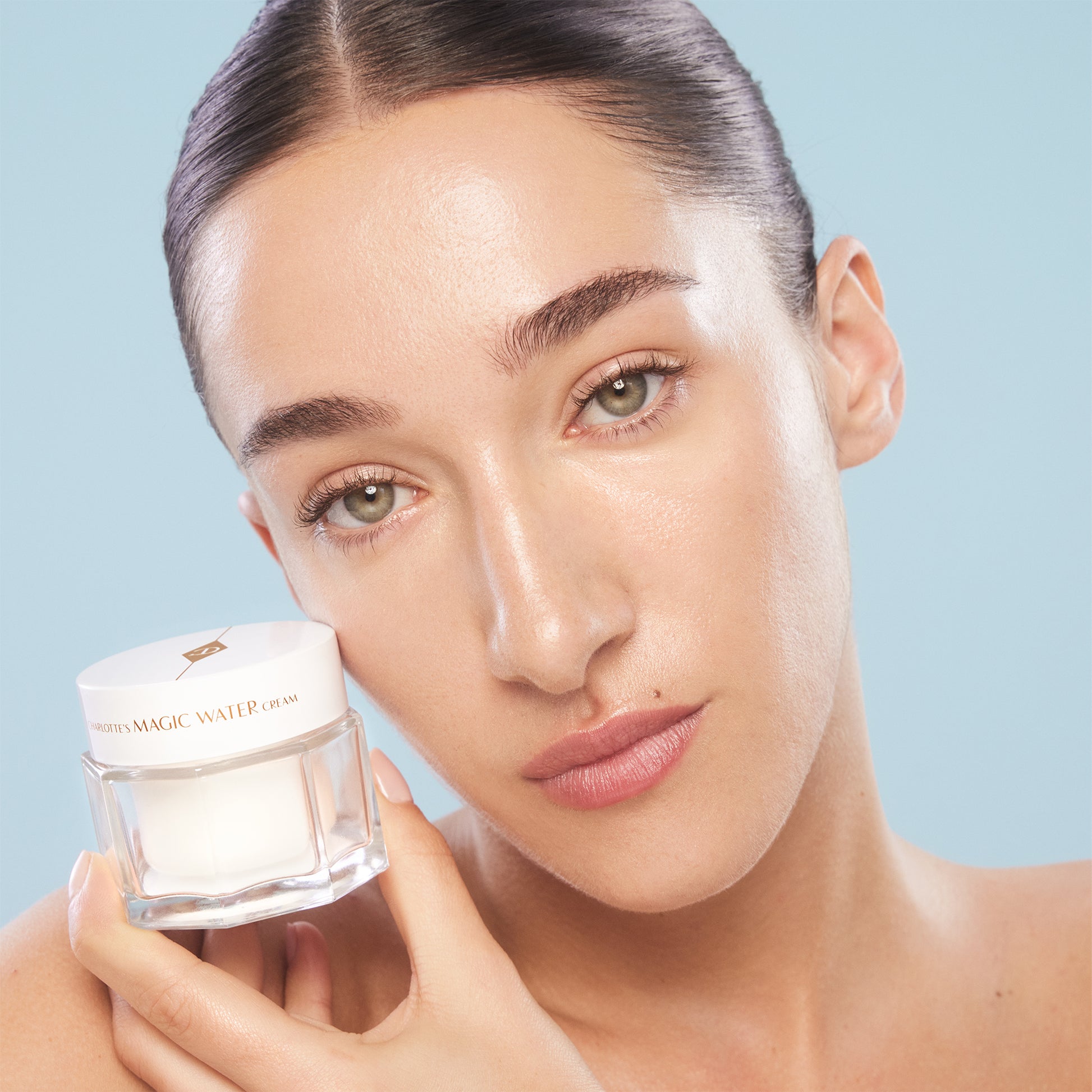 Model with glowing skin holding Charlotte Tilbury Magic Water Cream jar close to her face, at ZARÉ Cosmetics